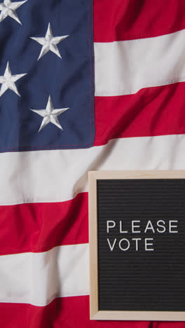 Vertical-Video-Of-Sign-Reading-Please-Vote-Lying-On-US-Stars-And-Stripes-Flag-For-American-Election-2024